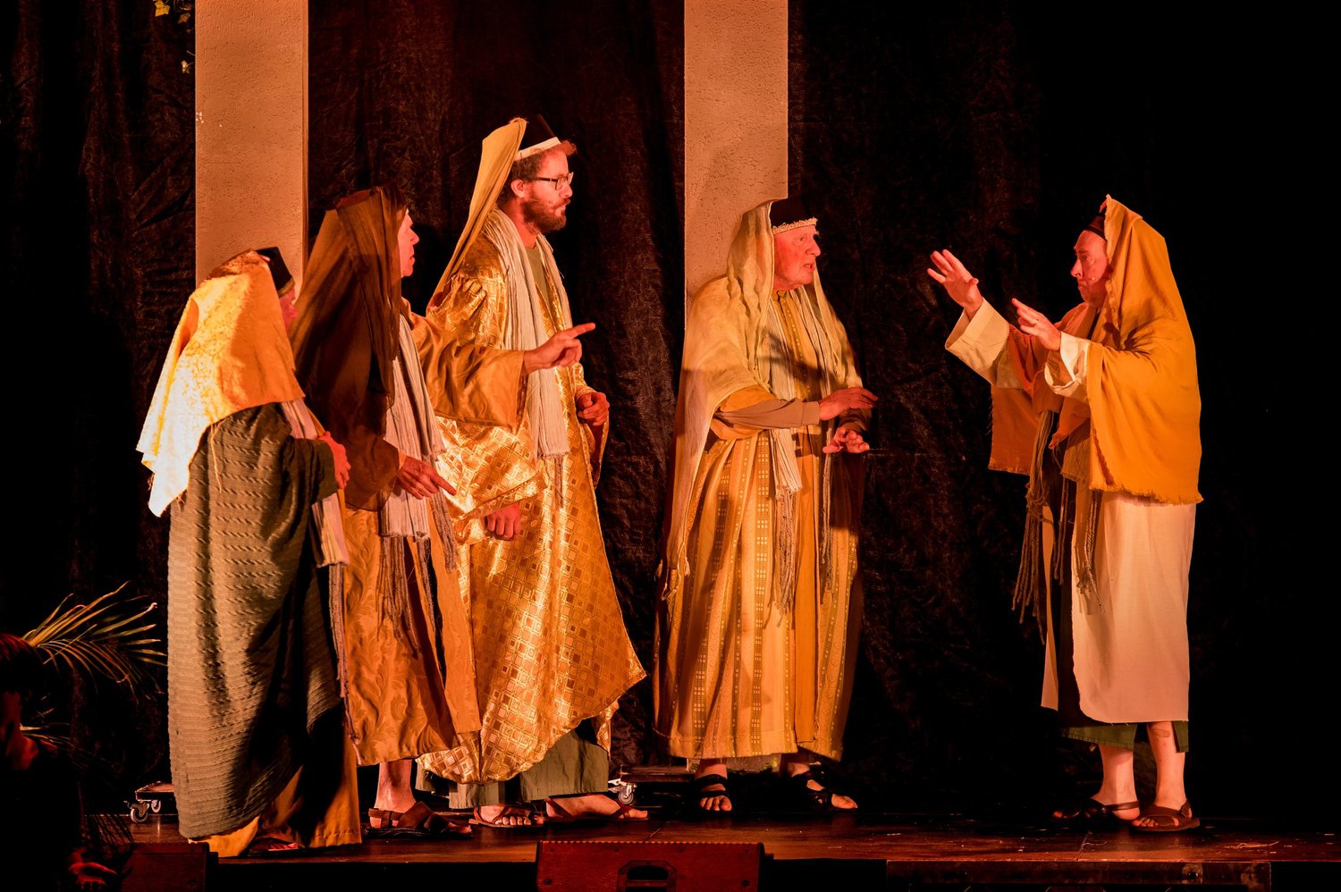 Priests in gold robes argue under warm stage light
